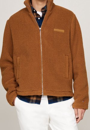 Brązowa bluza polarowa na zamek z bocznymi kieszeniami, noszona na niebiesko-brązowej koszuli w kratę i białych spodniach.