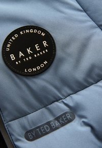 Niebieska tkanina o gładkiej fakturze, z czarną okrągłą metką z napisem "BAKER by TED BAKER" oraz szarym tagiem z napisem "BY TED BAKER."