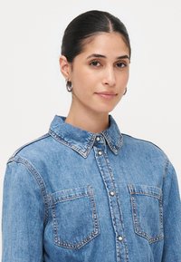 Camisa de ganga em azul claro, com colarinho pontudo, dois bolsos no peito, fecho com botões e detalhes de costura visíveis. Textura suave.
