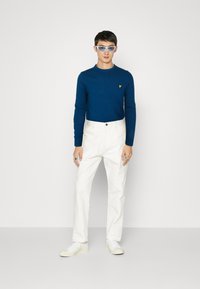 Maglietta a maniche lunghe a coste blu con logo giallo, pantaloni bianchi a gamba dritta e sneakers bianche, indossati con occhiali da sole celesti.