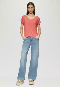 Korallenfarbenes V-Ausschnitt-T-Shirt mit umgeschlagenen kurzen Ärmeln, kombiniert mit hellblauen Weitbein-Jeans. Das Model steht mit Händen in den Taschen und trägt Plateausandalen.