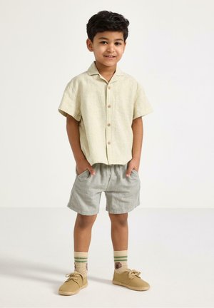 Ung dreng står med hænderne i lommerne på grå shorts, iført en beige kortærmet knappet skjorte, strømper og tanfarvede sko på hvid baggrund.