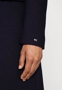 Tessuto di un cappotto blu scuro con una finitura testurizzata, caratterizzato da un piccolo logo dorato e rosso sull'orlo. Mano posizionata sulla manica.
