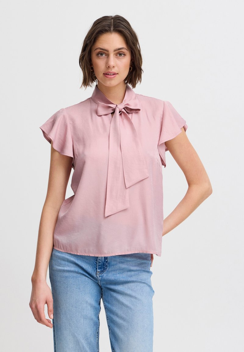 Blusa rosa chiaro a maniche corte con un fiocco legato al colletto, caratterizzata da una consistenza liscia e una vestibilità ampia. Indossata con jeans blu.