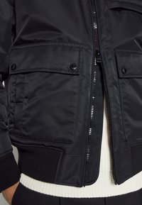 Svart bomberjacka tillverkad av glänsande nylon med två stora framfickor, dragkedjeförslutning och ribbade ärmslut. Har en kontrasterande vit innerfoder.