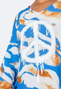 Top blu e bianco con motivo, caratterizzato da un grande simbolo della pace bianco con vernice che gocciola e dettagli in strass. Design a V, maniche lunghe.