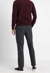 Burgunderfarbener Strickpullover kombiniert mit grauen Karohosen, die eine maßgeschneiderte Passform bieten. Schwarze Sneaker runden das Ensemble ab.