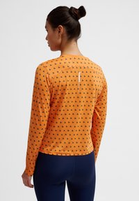 Langemouwen top in oranje met een herhaald zwart patroon. Heeft een ronde hals en een reflecterend logo op de achterkant. Glad, rekbaar materiaal.