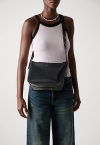 Zwarte leren schoudertas met een gladde textuur, hoekige vorm en metallic hardware-accenten; gedragen over een lichte tanktop en blauwe jeans.