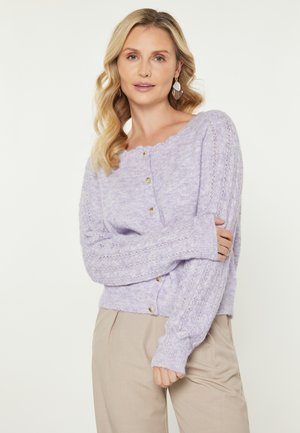 Lila neulecardigan, jossa on aaltoileva pääntie, napitus edessä ja teksturoitu kuvio hihoissa. Yhdistetty beiget housut.