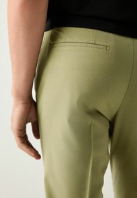Olijfgroen op maat gemaakte broek met een gladde textuur, voorzien van een enkele achterzak en een slimfit silhouet. Hand rustend naast het kledingstuk.