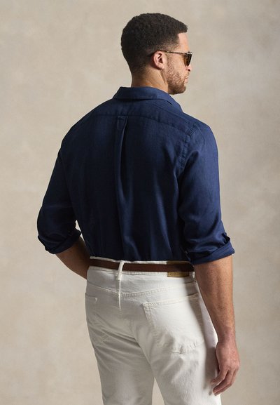 Jūras zils lina krekls ar sarullētām piedurknēm, kombinēts ar baltām biksēm un brūnu jostu, skatīts no aizmugures. Ar teksturētu audumu un pielāgotu griezumu.