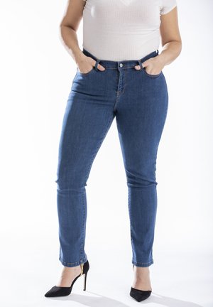 Jeans en denim blue slim-fit avec taille mi-haute, poches avant et fermeture à bouton. Associés à des escarpins noirs à talons aiguilles sur un fond blanc.