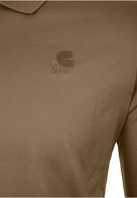 Beige Poloshirt mit einem kleinen gestickten Silhouettenlogo eines bärtigen Mannes auf der linken Brust.