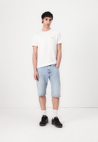 T-shirt blanc à manches courtes avec un petit logo, shorts en denim bleu clair et baskets noires avec des chaussettes blanches. Tenue simple et décontractée.