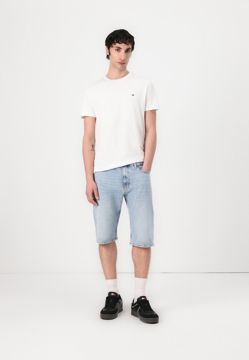 T-shirt blanc à manches courtes avec un petit logo, shorts en denim bleu clair et baskets noires avec des chaussettes blanches. Tenue simple et décontractée.