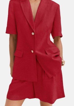 Kobieta w dopasowanym czerwonym marynarce z krótkim rękawem z guzikami i pasujących czerwonych szortach z wysokim stanem, ręka w kieszeni.