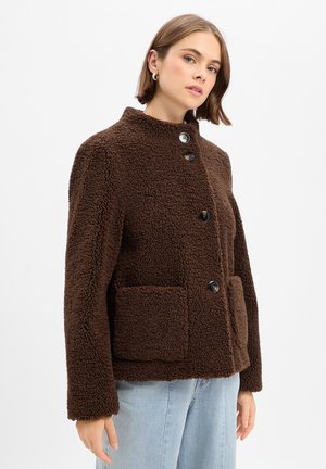 Junge Frau trägt eine braune, strukturierte Jacke mit Knöpfen und großen Fronttaschen sowie hellblaue Jeans vor einem einfarbigen Hintergrund.