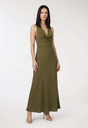 Donna con capelli lisci e scuri che indossa un abito lungo senza maniche verde oliva e sandali con tacco alto dorati e a listini, in piedi davanti a uno sfondo semplice.