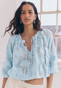 Blusa corta azzurro chiaro con ricami floreali, scollo arricciato con chiusure a nodo e maniche larghe a tre quarti.