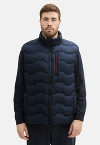 Gilet doudoune bleu marine avec un col haut, présentant une fermeture éclair à l'avant, un motif matelassé horizontal et une poche poitrine avec des accents rouges.
