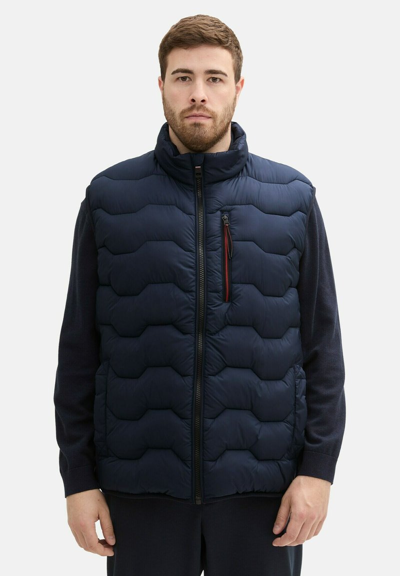 Gilet doudoune bleu marine avec un col haut, présentant une fermeture éclair à l'avant, un motif matelassé horizontal et une poche poitrine avec des accents rouges.