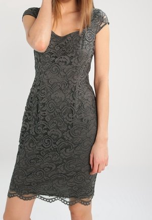 Kvinde iført en tætsiddende, knælængde mørkegrå blondekjole med kappableder og en bølget kant, der står mod en ensfarvet baggrund.