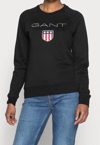Sudadera negra con cuello redondo y mangas raglán largas. La parte frontal presenta el logo "GANT" con un escudo y acentos en rojo y azul. Textura suave.