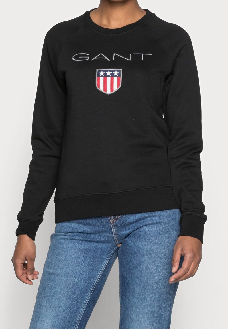 Sudadera negra con cuello redondo y mangas raglán largas. La parte frontal presenta el logo "GANT" con un escudo y acentos en rojo y azul. Textura suave.