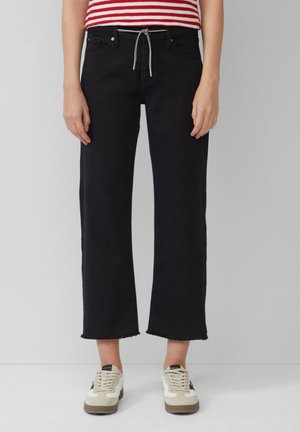 Schwarze weit geschnittene Jeans mit rohem Saum, ausgestattet mit einem Kordelzugbund, Vordertaschen und getragen mit beigefarbenen und schwarzen Sneakers.