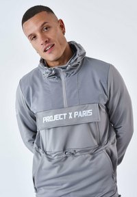 Giacca a hoodie grigia con zip frontale, dotata di un grande pannello orizzontale etichettato "PROJECT X PARIS" in bianco. Materiale liscio e leggero.