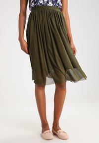 Femme portant une jupe midi plissée olive, un haut floral bleu marine et des mocassins rose clair, se tenant devant un fond blanc uni.