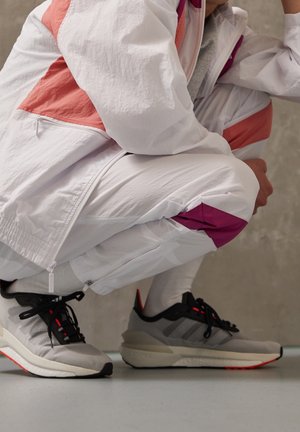 Persoon in hurkzit, gekleed in witte en roze sportkleding met grijze en zwarte sneakers, op een lichte vloer tegen een grijze muur.