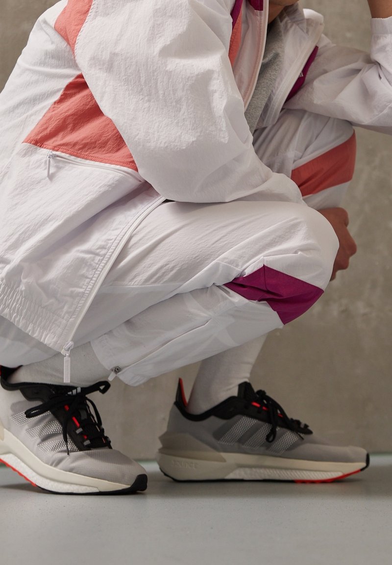 Personne accroupie portant une tenue de sport blanche et rose avec des baskets grises et noires sur un sol clair contre un mur gris.