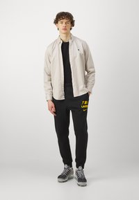 Lys grå knapp-up skjorte over en svart T-skjorte, parret med svarte joggebukser med gule "Lakers"-tekst. Grå og svarte sneakers.