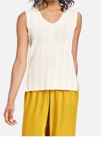 Beżowa, bezrękawnikowa bluzka o prążkowanej strukturze z dekoltem w kształcie litery V, w zestawieniu z luźnymi, musztardowymi spodniami, na stojącej osobie o zrelaksowanej postawie.