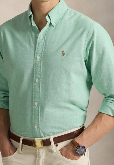 Ανοιχτό πράσινο πουκάμισο με κουμπιά και μικρό, κεντημένο λογότυπο αλόγου. Συνδυασμένο με καφέ δερμάτινη ζώνη και μπλε ρολόι στον καρπό.
