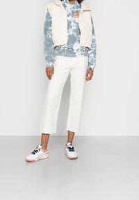 Kremowa, pikowana kamizelka noszona na niebieskiej bluzie z kapturem w stylu tie-dye, zestawiona z białymi, krótkimi spodniami i wielokolorowymi sneakersami z różowymi i czarnymi akcentami.