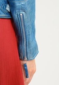 Nahaufnahme eines blauen Lederjackenärmel mit einem silbernen Reißverschluss, der über einem roten Stoffkleidungsstück getragen wird, wobei das Handgelenk und die Hand sichtbar sind.