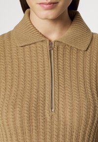 Beige stickad tröja med dragkedja vid kragen, med ett texturerat ribbmönster. Tyget är mjukt med synliga s ömmar och en subtil lyster.