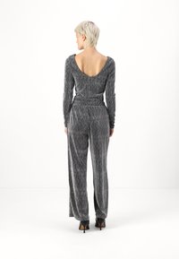 Combinaison en argent métallique avec manches longues et décolleté profond dans le dos, présentant une taille cintrée et un pantalon large. Texture lisse et design ajusté.