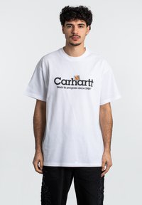 Camiseta de algodón blanca con el logotipo negro de "Carhartt" y el texto "Trabajo en progreso desde 1989". Mangas cortas y un ajuste relajado.