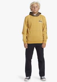 Gul hoodie med bruna detaljer, framficka för känguru och logotypdetalj. Bärs med svarta byxor och svart-vita sneakers.