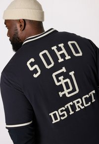 Námořnická modrá košile na knoflíky s bílým vyšívaným textem "SOHO" a "DSTRCT" na zadní straně. Obsahuje žebrovaný límec s bílými pruhy.