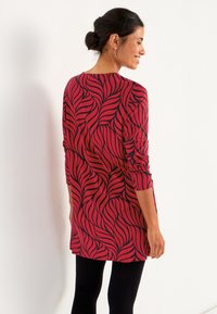 Langärmlige Tunika in Rot mit einem schwarzen Blattmuster, mit lockerem Schnitt und rundem Ausschnitt, kombiniert mit schwarzen Leggings.