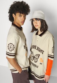 Beige polo skjorter med brune detaljer og grafikk, med sommerfugler og tekst. En modell har på seg et strukturet oransje langermet lag.