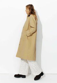 Manteau long beige avec un design à boutons sur le devant, des poches latérales et une coupe décontractée. Porté avec un pantalon blanc à motifs et des bottes chelsea marron foncé.