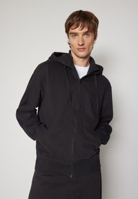 Sudadera con cremallera negra hecha de tela suave, con un bolsillo tipo canguro, capucha con cordón y puños acanalados. El modelo se encuentra de pie con las manos en los bolsillos.