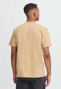 Lyhythihainen, pehmeästä puuvillasta valmistettu tan väri t-paita. Crew neck -muotoilu, jossa on minimaalinen saumasuunnittelu, yhdistettynä tummiin farkkuihin ja näkyviin ompeluseikkoihin.