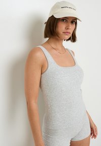 Body in cotone grigio con scollatura quadrata e texture a costine, abbinato a un cappellino crema con testo nero, indossato con una semplice collana.